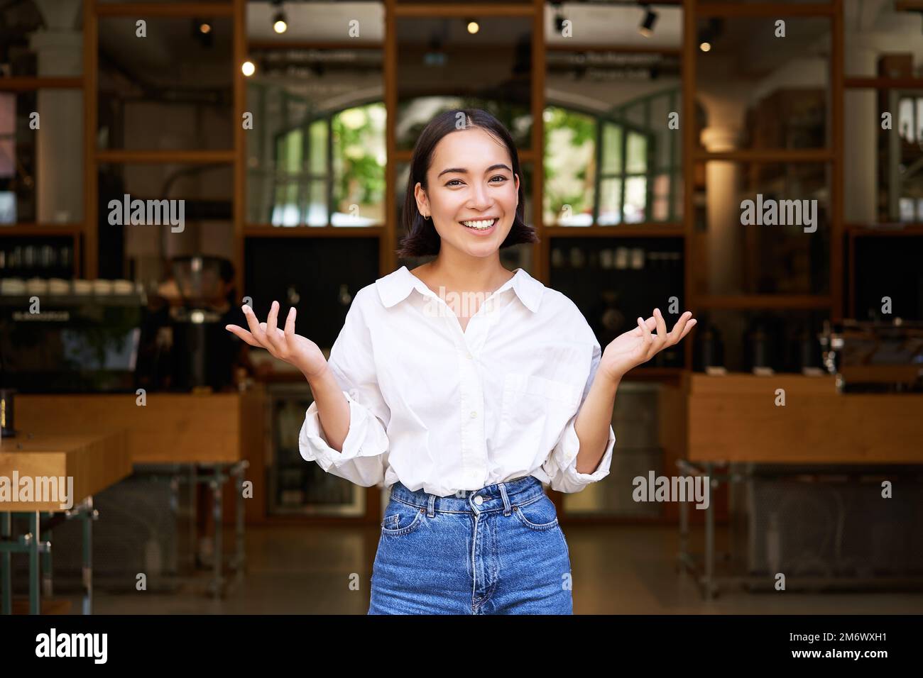 Enthusiastic young businesswoman, asian girl showing her business, raising hands up and smiling, standing in front of restaurant Stock Photo