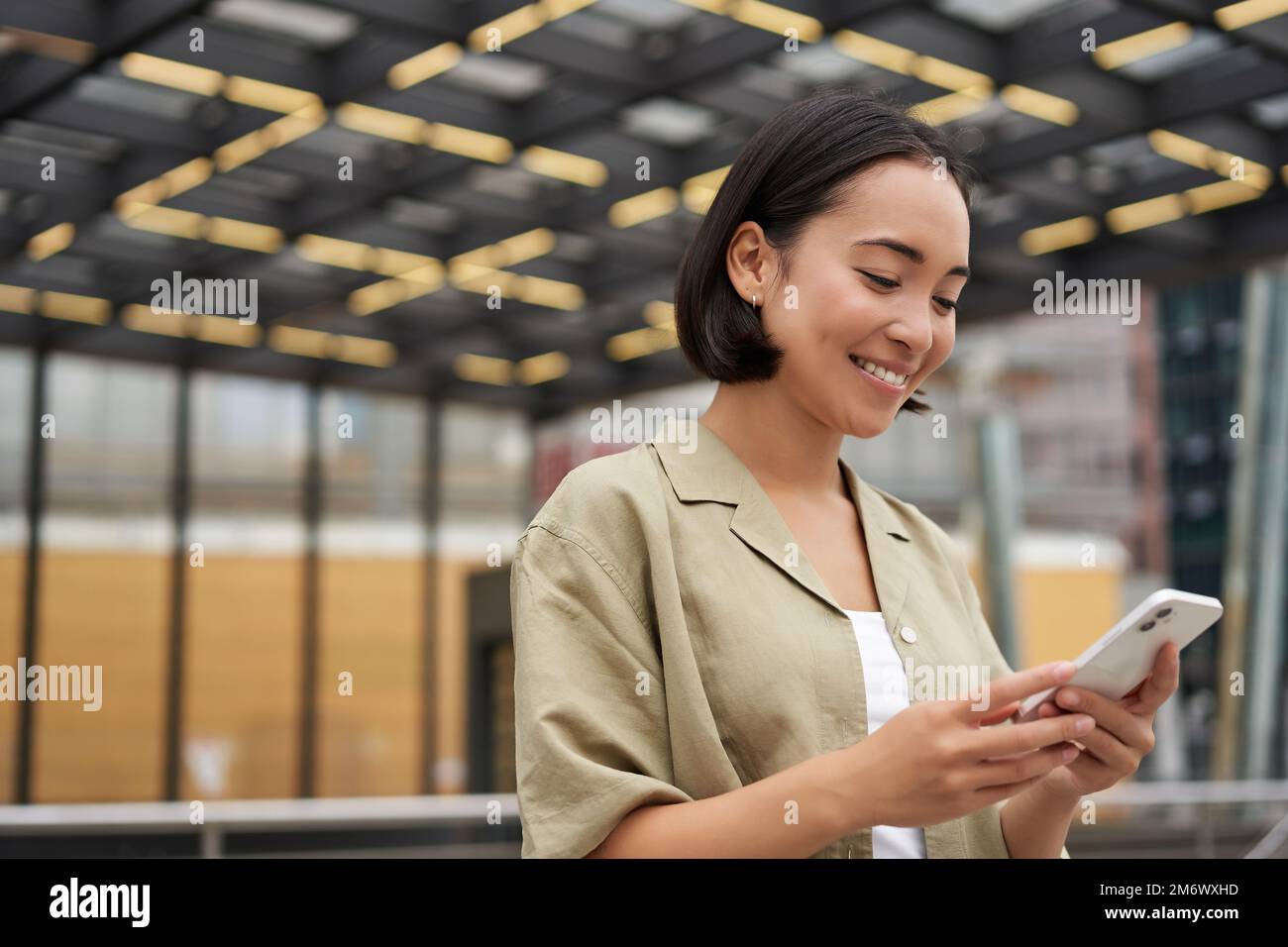 Technology people. Young smiling asian girl holding smartphone, using mobile app while standing on street, navigating around cit Stock Photo