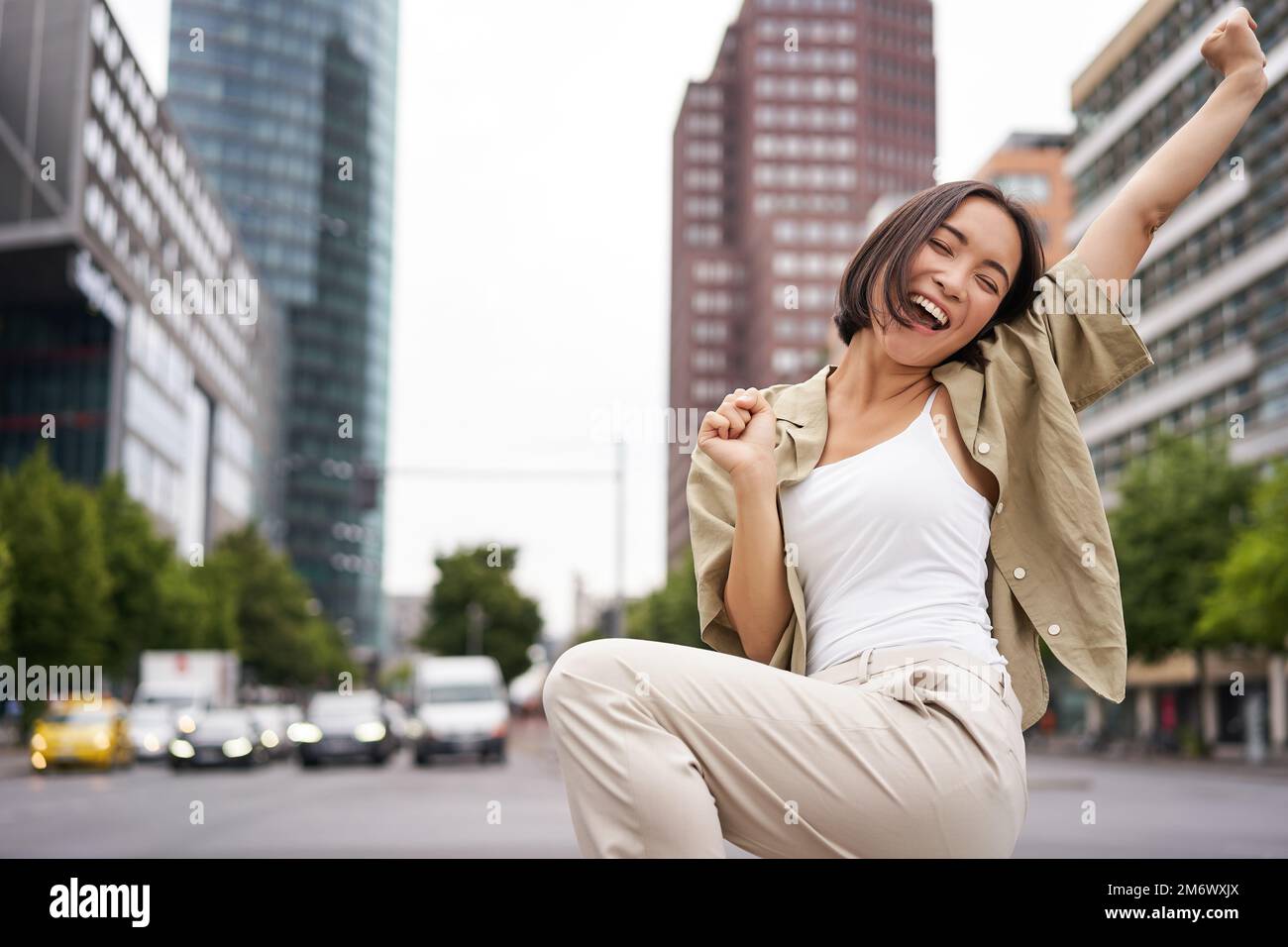 Portrait of happy asian woman, dancing and feeling joy, triumphing, raising hand up in victory gesture, celebrating on streets Stock Photo