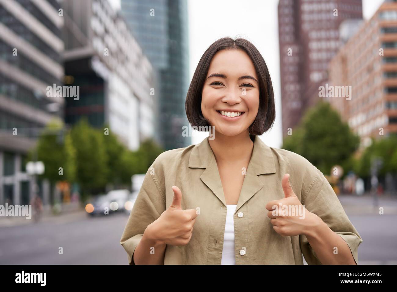 Enthusiastic asian woman , shows thumbs up in approval, looking upbeat, say yes, approves and agrees, stands on street Stock Photo