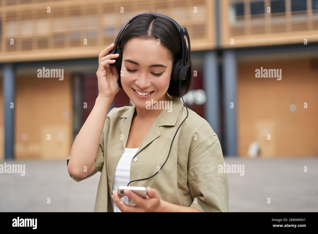 Streaming services concept. Happy asian girl listens music in headphones, holds mobile phone, choosing track or podcast, walking Stock Photo