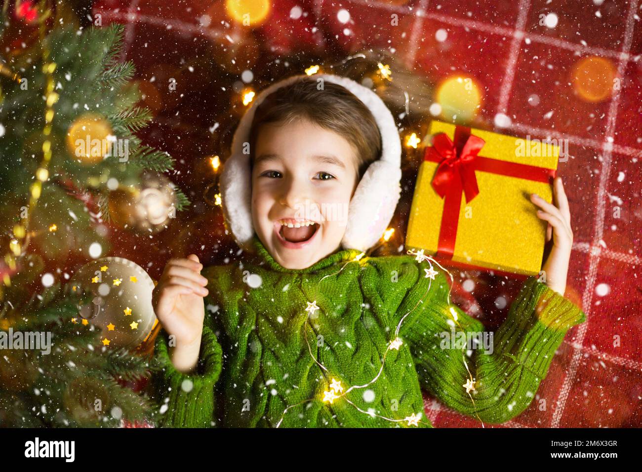 Girl in a warm knitted sweater and fur headphones is lying on a blanket near the Christmas tree with a gift box. Christmas Eve, Stock Photo