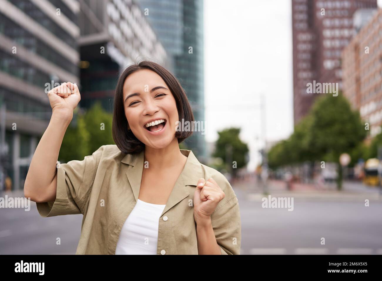 Happy asian girl winning and rejoicing, celebrating on streets of city, dancing from joy, triumphing and smiling Stock Photo