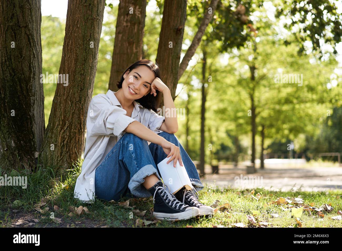 Woman sitting in park with her favourite book, leaning on tree under shade on sunny day, enjoying nature and calm relaxing atmos Stock Photo