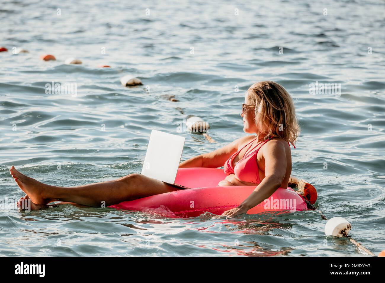Woman works on laptop in sea. Freelancer, young blond woman in sunglases floating on an inflatable big pink donut with a laptop in the sea at sunset Stock Photo