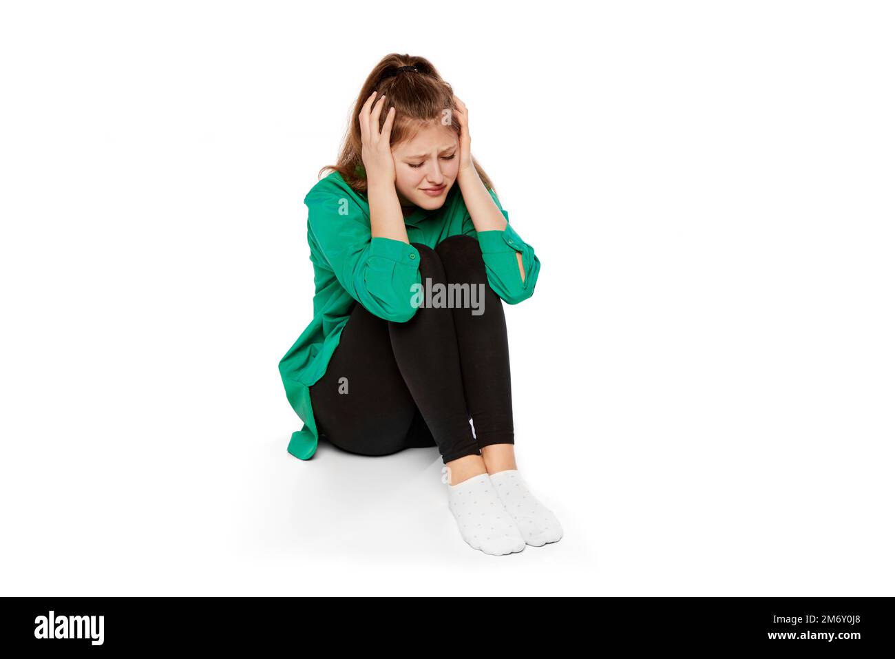 Portrait of depressed teen girl sitting on floor and covering ears with hands over white background. Breakdown, apation Stock Photo