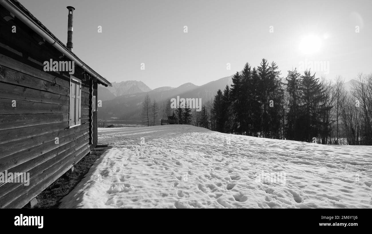 Wooden hut with shutters and narrow chimney in the snow where footsteps can be seen and sunrays, forest and mountains in background in black and white Stock Photo