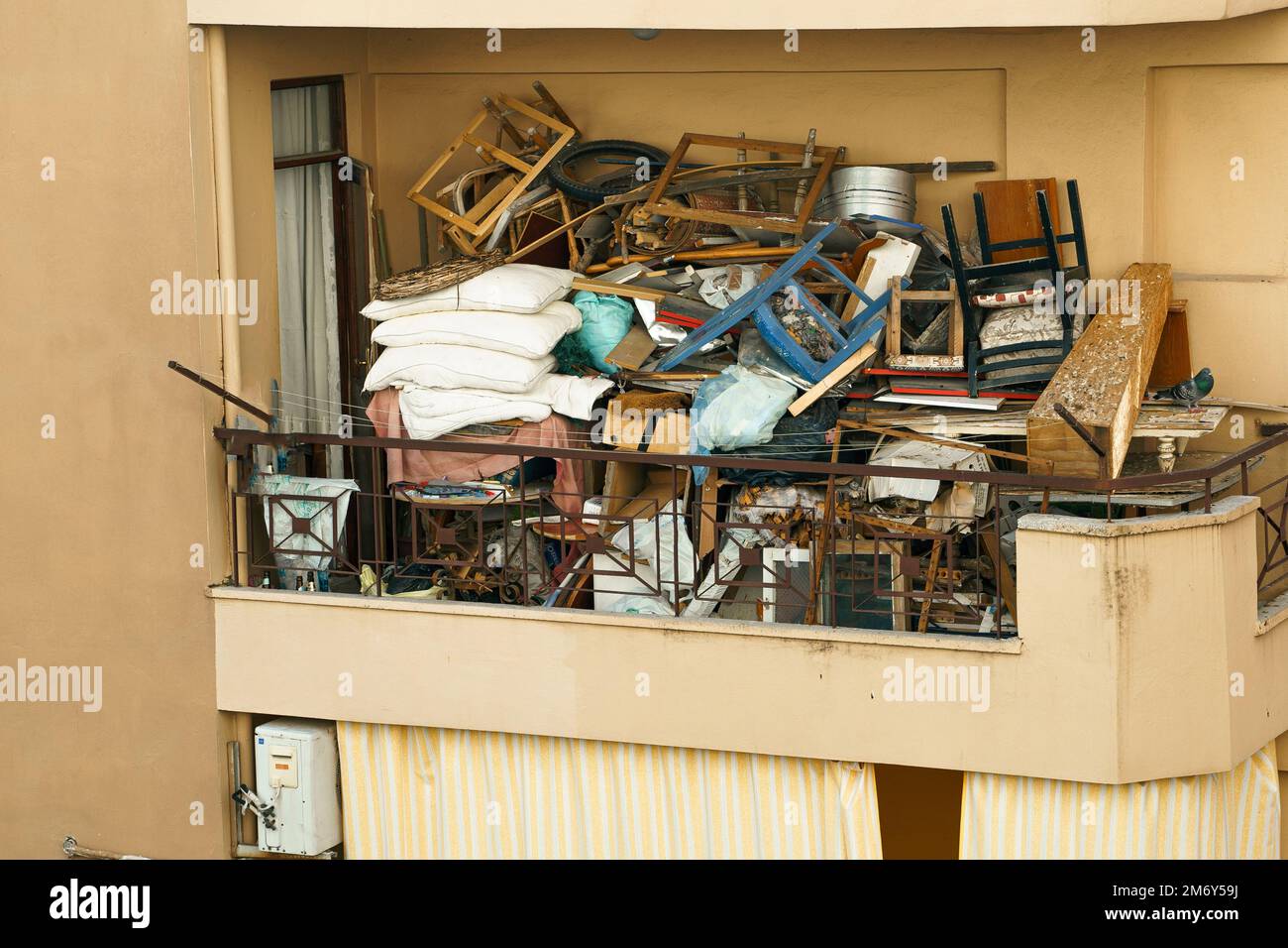 Ugly, terrible balcony disorder. Multi-storey building full of garbage, old furniture, unnecessary things, boxes,chairs, pillows, clothes, window fram Stock Photo