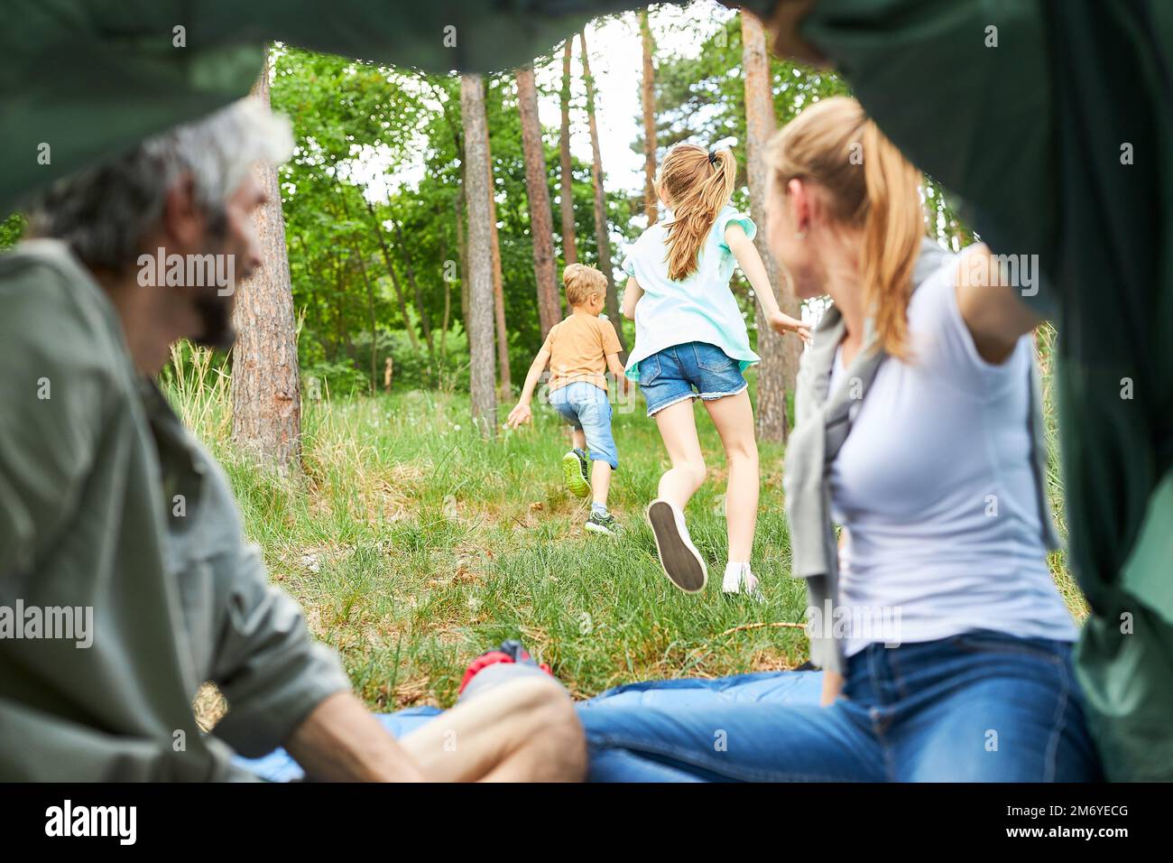 Mother and daughter sitting inside tent looking at playful children running in forest Stock Photo