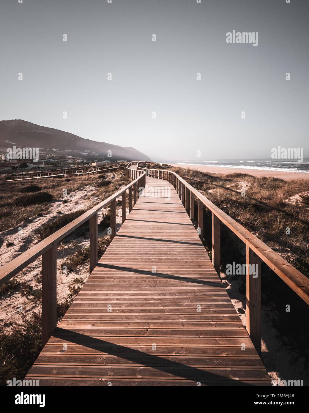 A long boardwalk on a grassy sand dune at the beach leading to the mountain and ocean with desaturated colors. The way ahead concept, Quiaios Beach Stock Photo