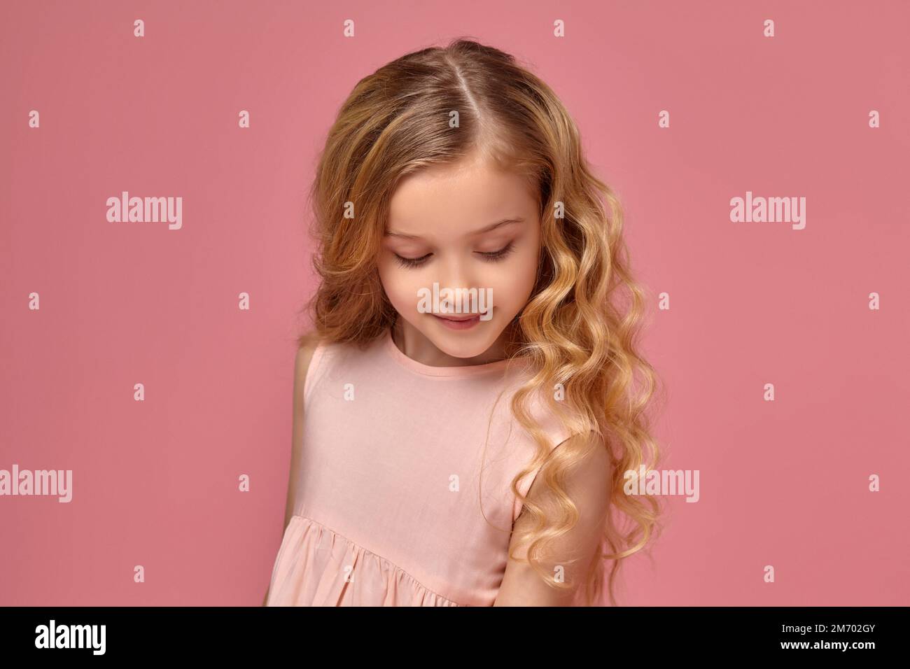 Little girl with a blond curly hair, in a pink dress is posing for the ...