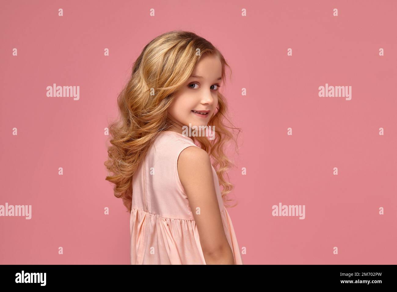 Little girl with a blond curly hair, in a pink dress is posing for the ...