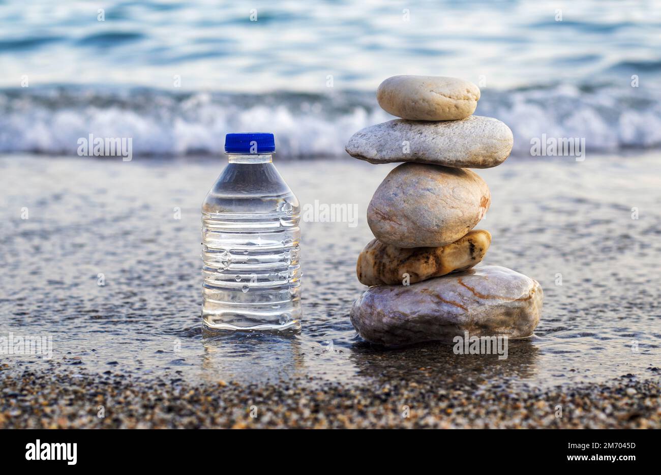 pebbles in pyramid one over another and bottle with drinking water on big rock beach sea waves in background.pure water.bottle and stones surrounded b Stock Photo