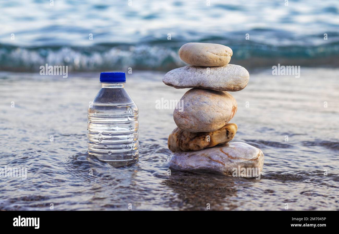 pebbles in pyramid one over another and bottle with drinking water on big rock beach sea waves in background.pure water.bottle and stones surrounded b Stock Photo