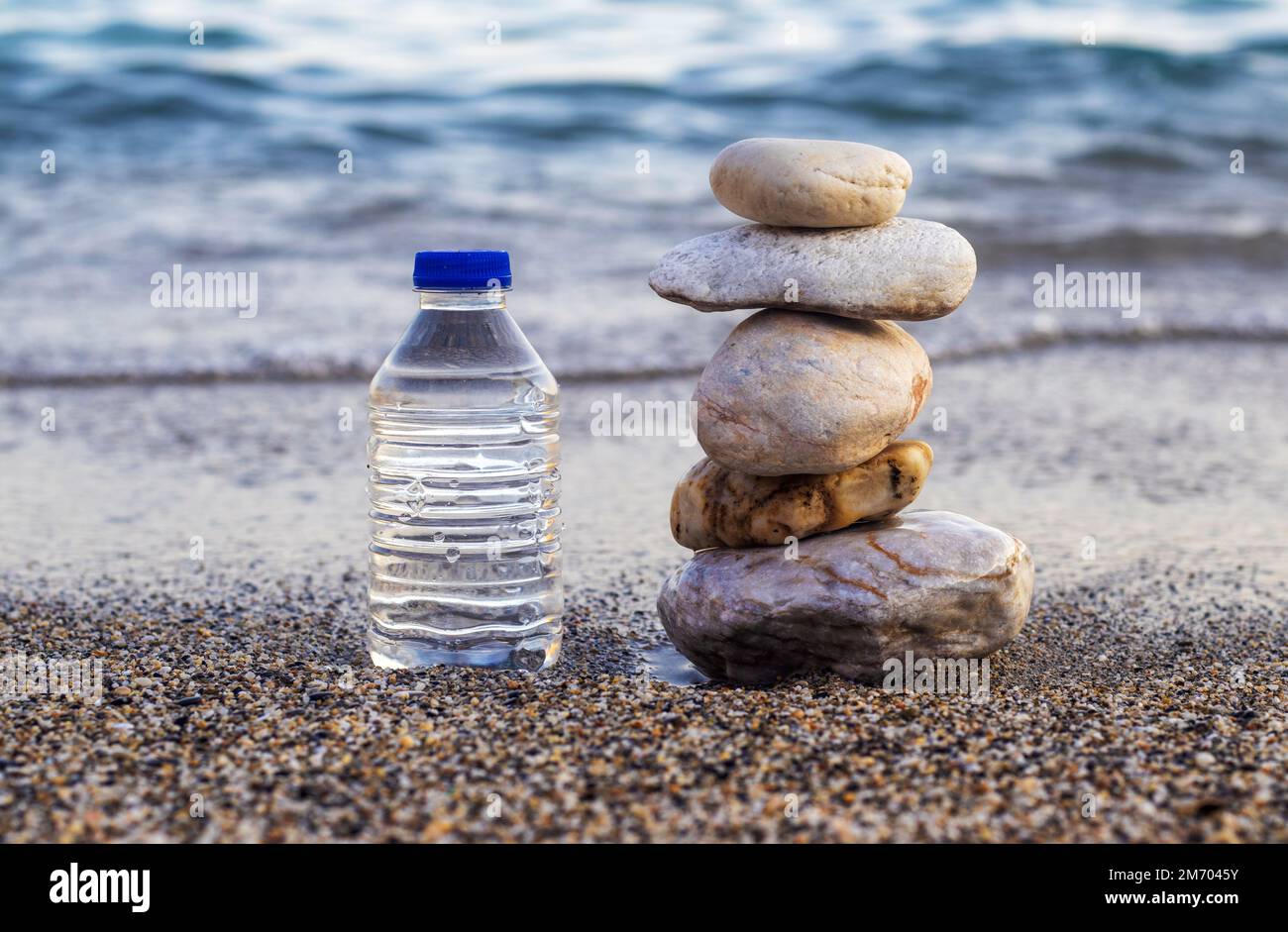 pebbles in pyramid one over another and bottle with drinking water on big rock beach sea waves in background.pure water.bottle and stones surrounded b Stock Photo