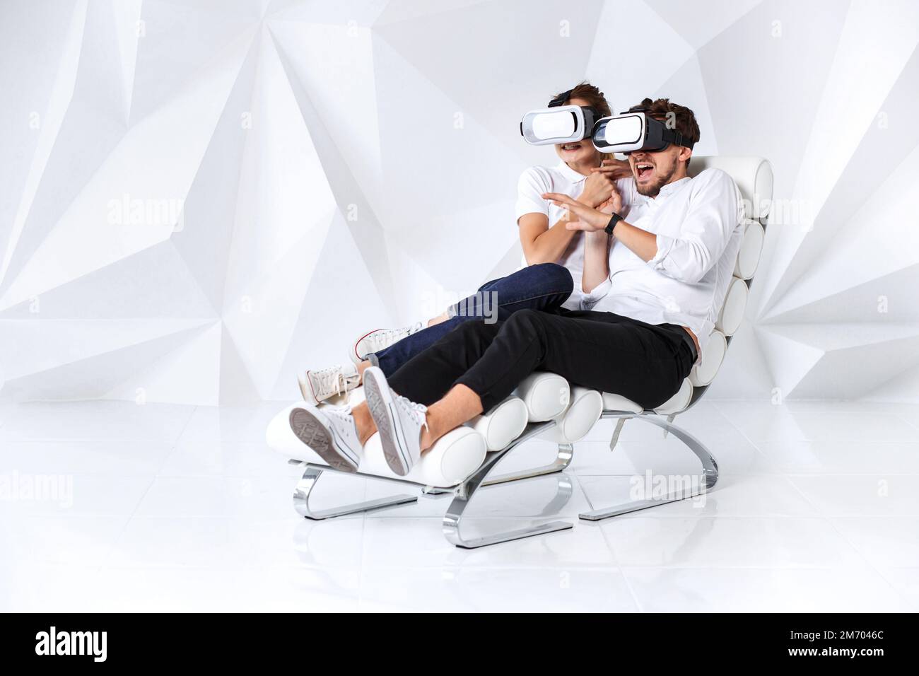 A young couple wearing VR headsets sitting on white chair in a room ...