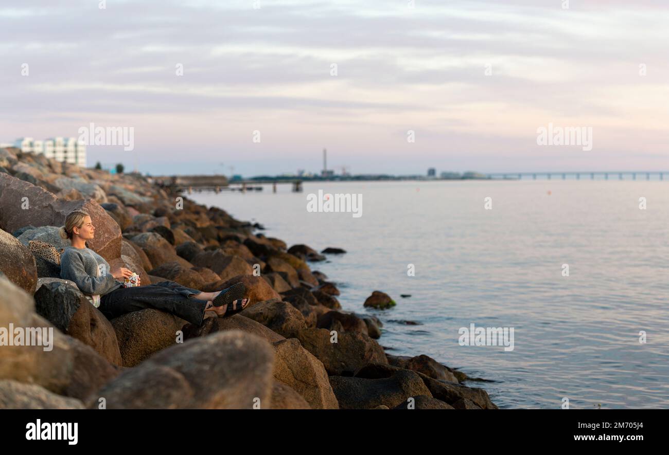 A young woman sitting on rock near the ocean eating popcorn while watching the sunset in south Sweden Stock Photo