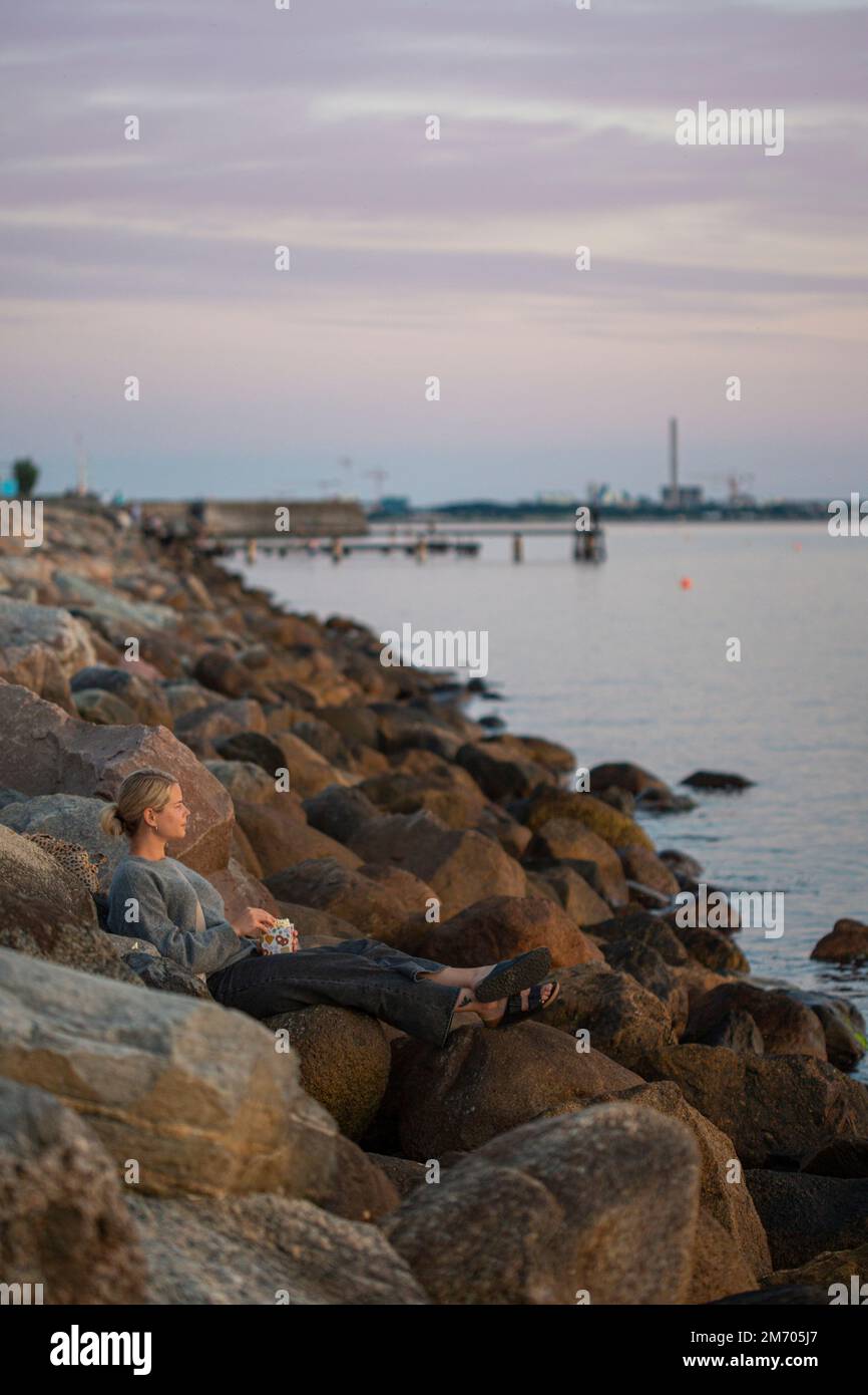 A young woman sitting on rock near the ocean eating popcorn while watching the sunset in south Sweden Stock Photo