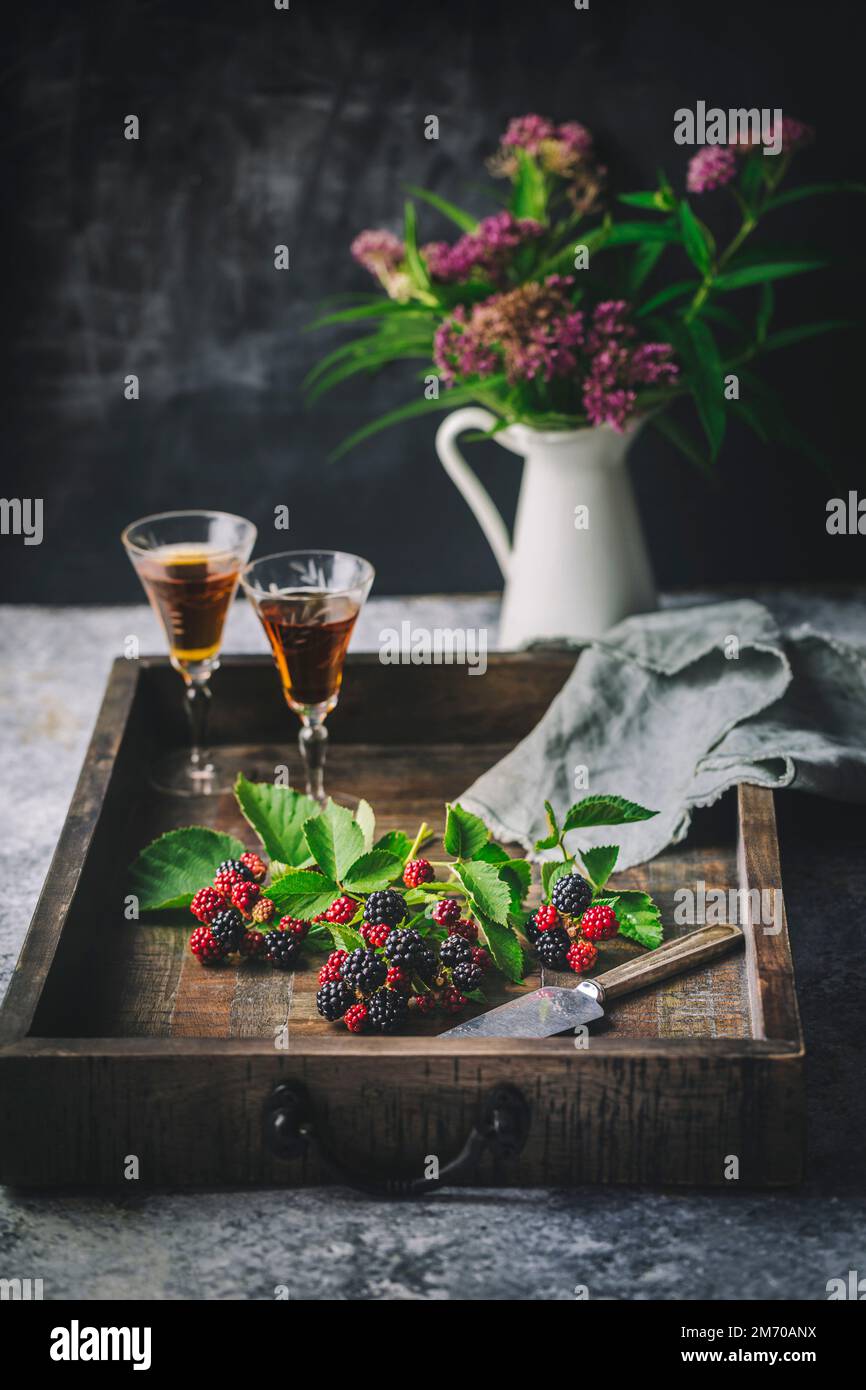 Blackberries and leaves on wooden tray with sherry in glasses and flowers in vase Stock Photo