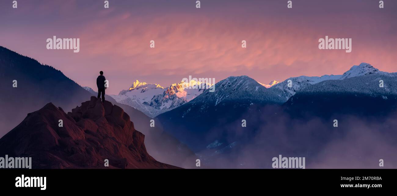 Adventurous Man Hiker standing on top of icy peak with rocky mountains in background. Stock Photo