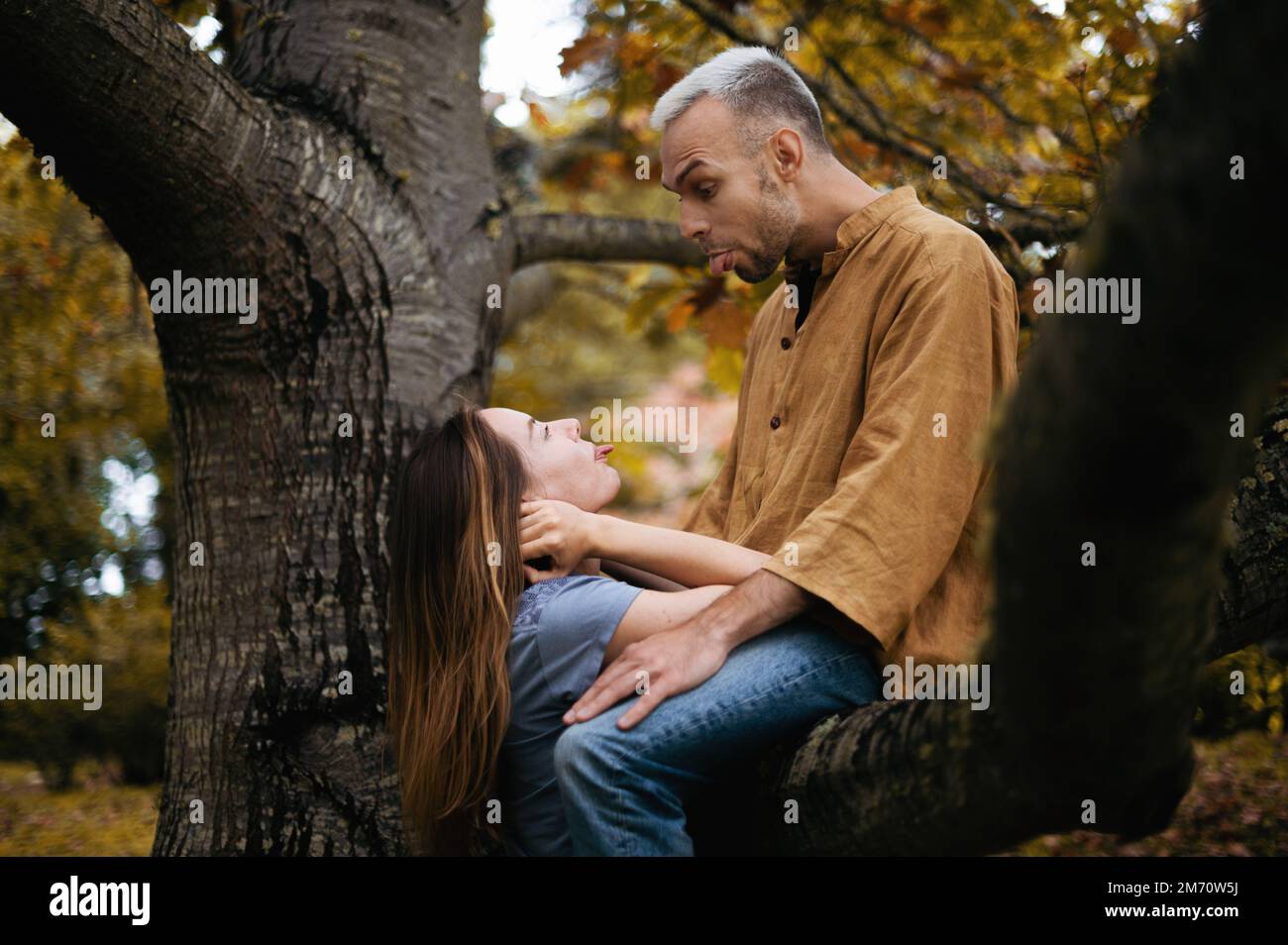 A playful and romantic photo of a young couple in a beautiful autumn park, with the man sitting on a tree Stock Photo