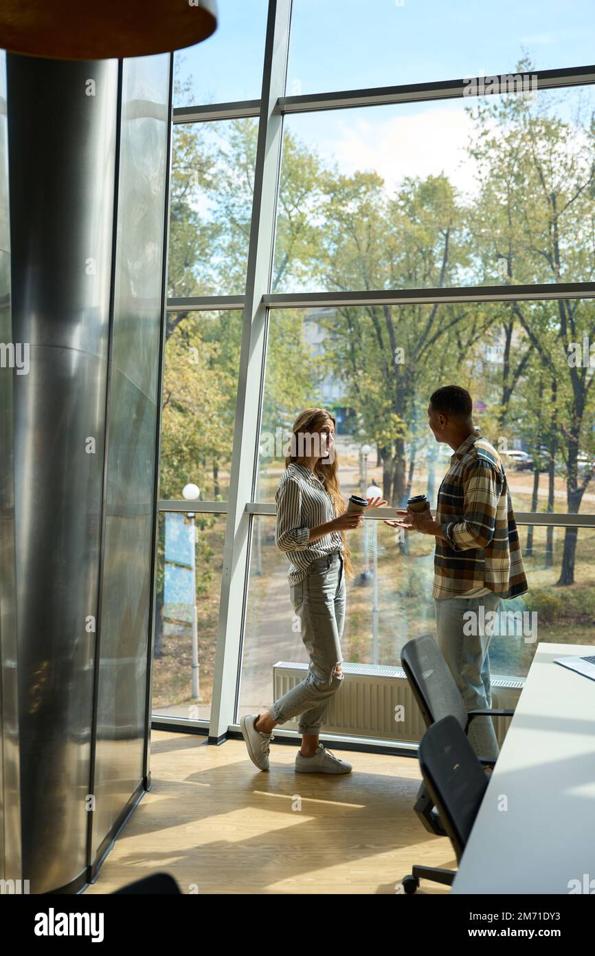 Two colleagues holding coffee while talking near big window Stock Photo