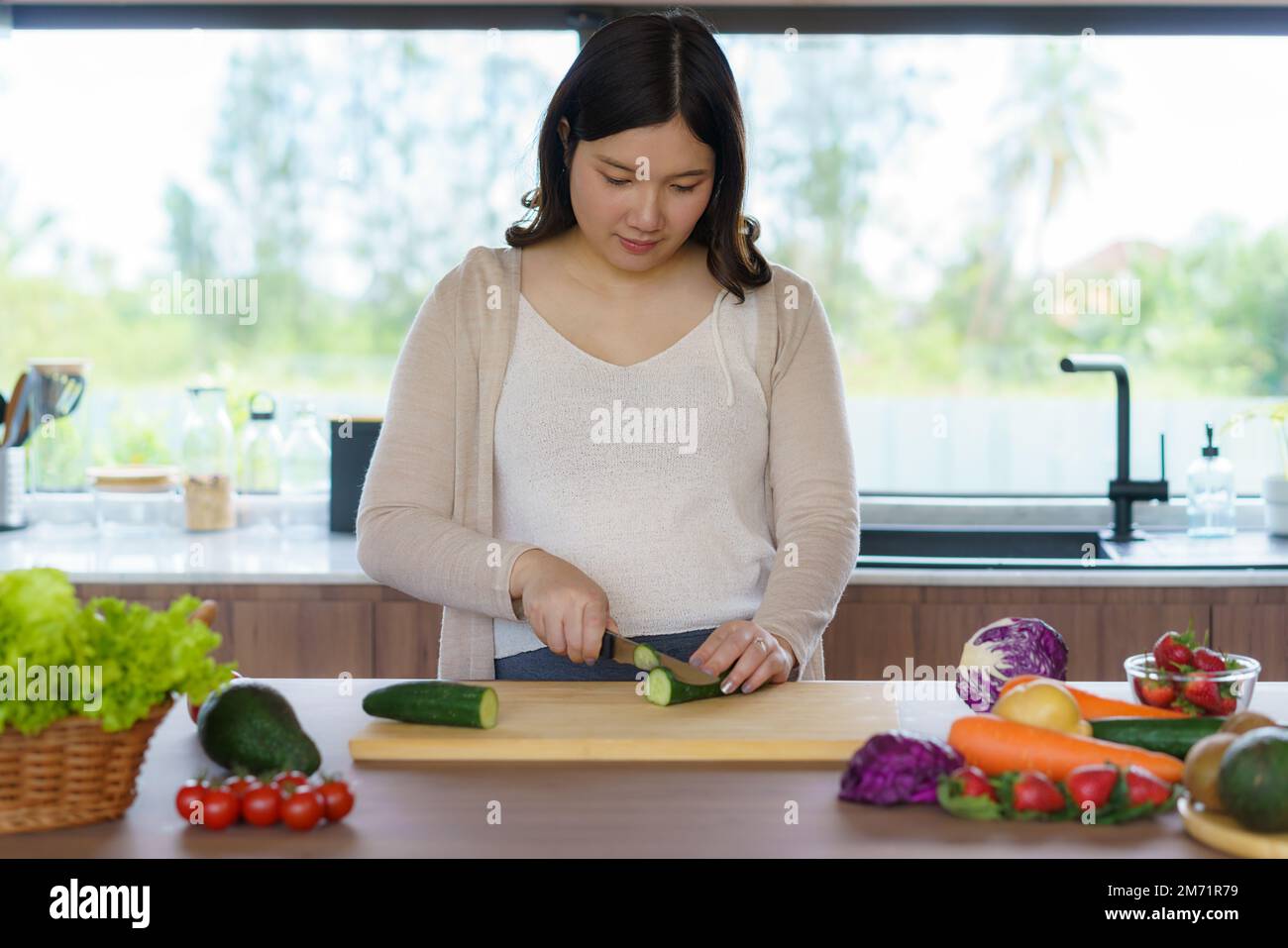 Pregnant Asian woman cutting cucumber for fresh green salad, female prepares tasty organic dinner at home, healthy nutrition for future mother Stock Photo