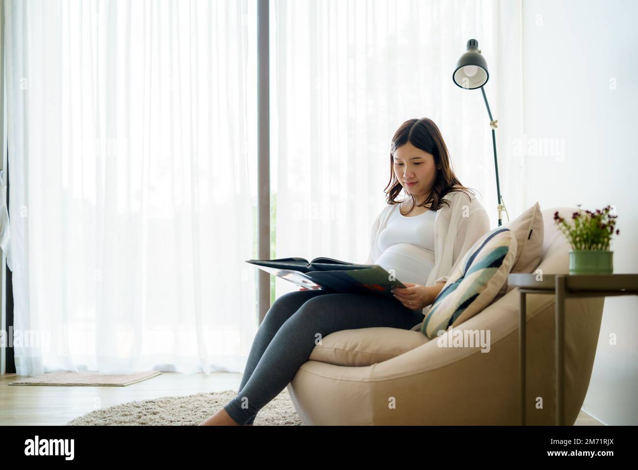 Asian Pregnant woman read a book for baby in belly. Mom feeling happy smiling positive and peaceful while take care child lying on sofa in living room Stock Photo