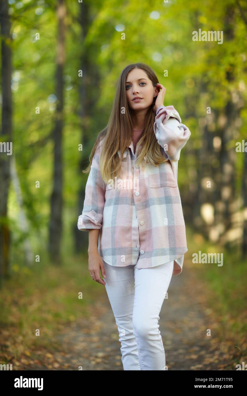 Blonde female in casual dress walking along autumn linden tree alley. Path under yellow trees with falling autumn leaves. Sunny autumn day. T Stock Photo