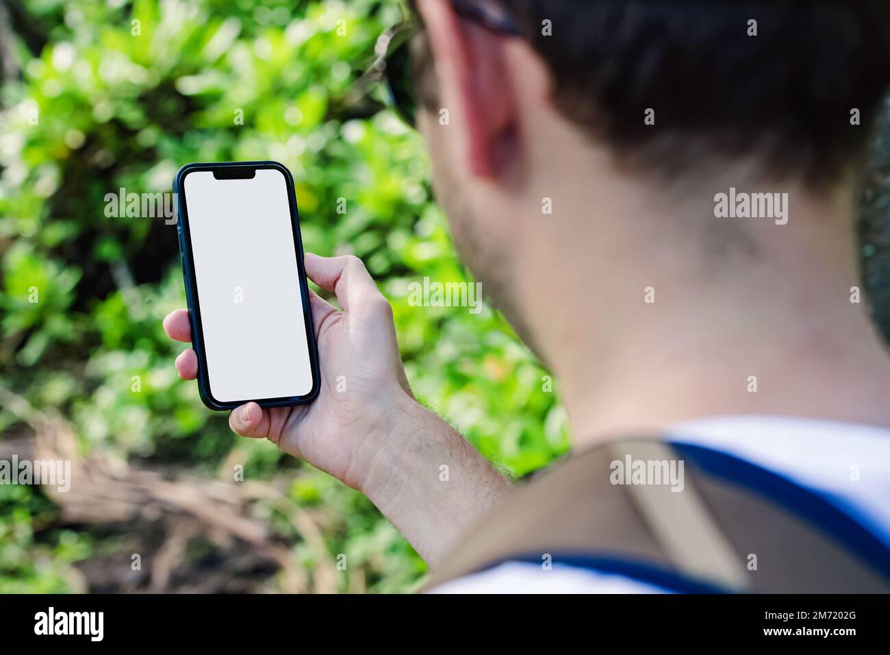Man holding a smart phone with a blank white screen on the background of blurred lush greenery. Phone mock-up Stock Photo