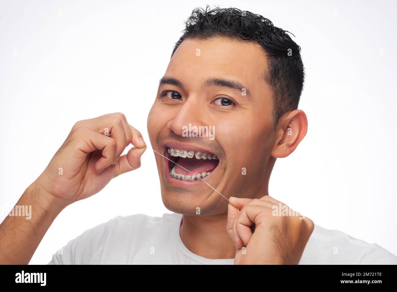 Hispanic man in T-shirt smiles as he flosses his teeth with orthodontic treatment brackets on a white background, one person Stock Photo
