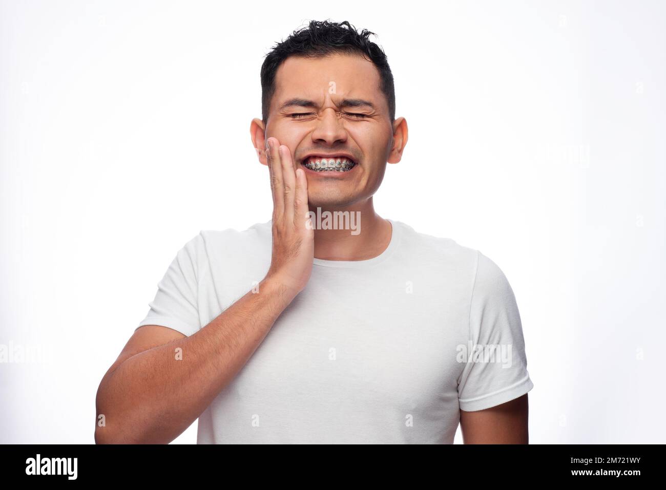 Hispanic man in white shirt and braces grimaces in pain on his face and holds his face by his orthodontic treatment brackets, one person Stock Photo