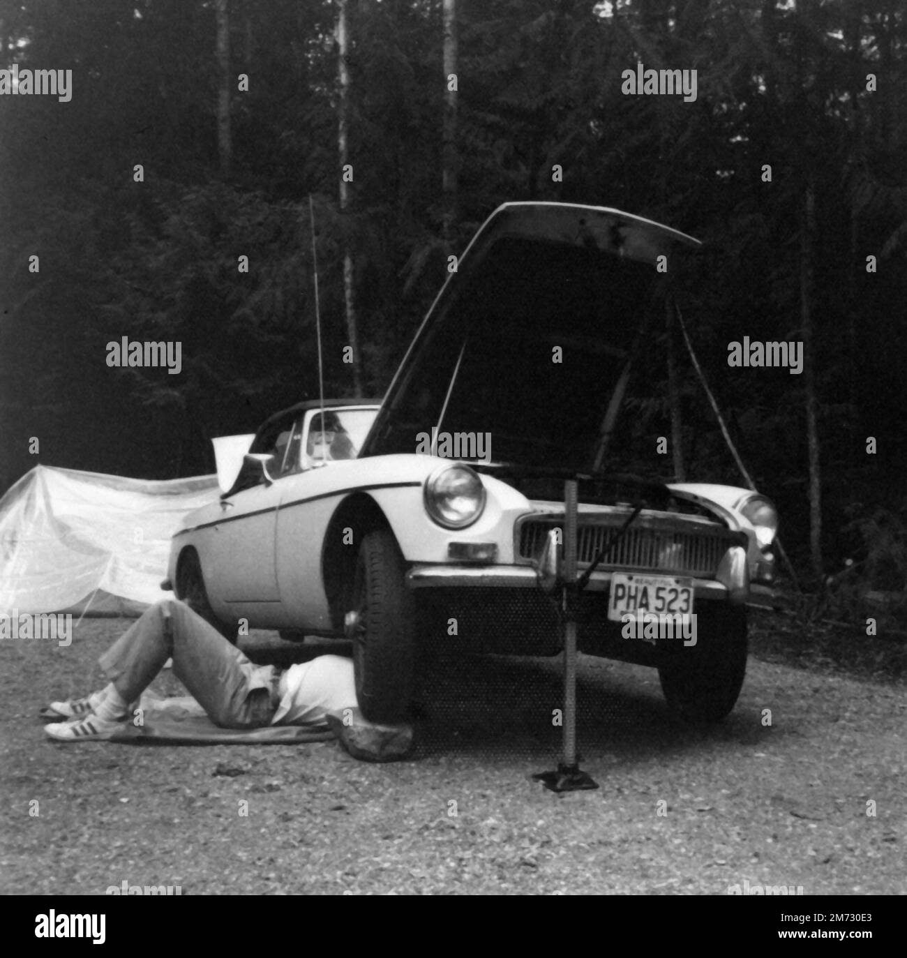 Man lying under a car to repair it. The car is a white MG MGB from 1968. Stock Photo