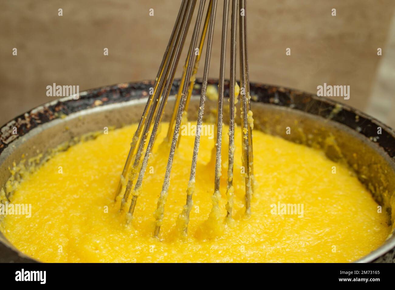 Boiling corn flour in a cast iron pot, making mamaliga, traditional romanian food, and mixing with a stainless steel whisk, soft focus close up Stock Photo