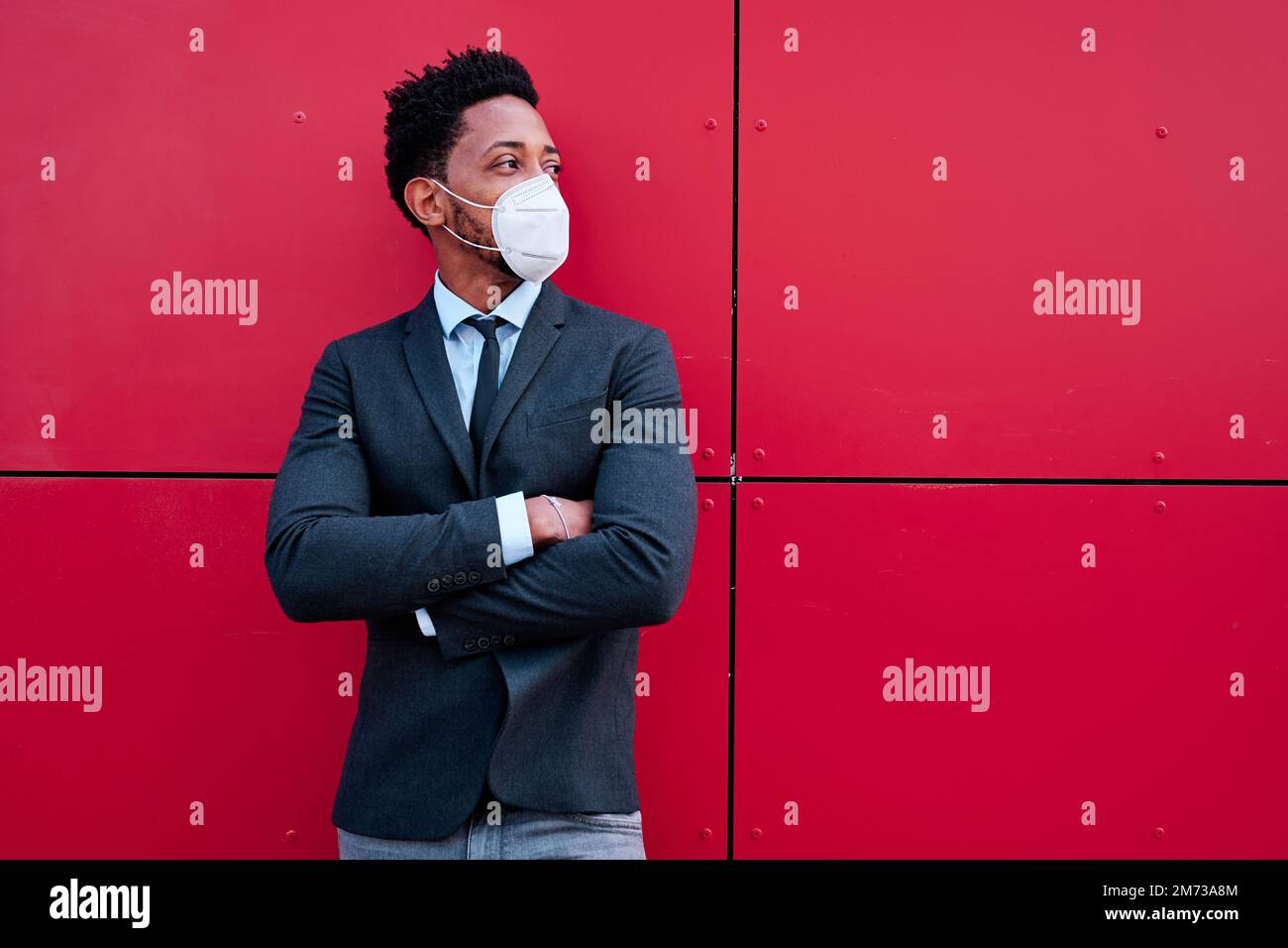 Male entrepreneur with face mask looking away while standing with arms crossed outdoors. Stock Photo