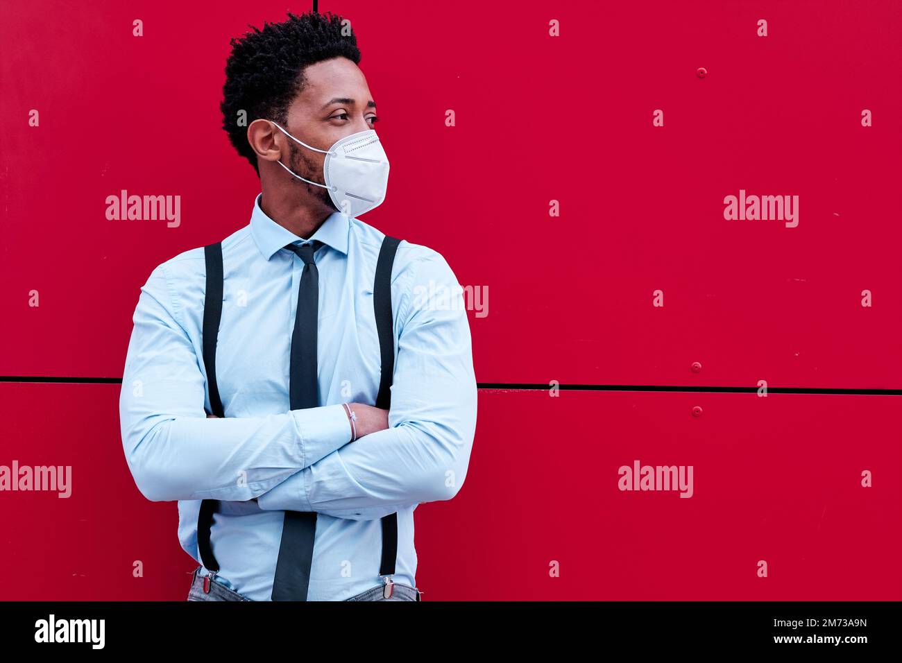 Businessman with a face mask looking away while standing with his arms crossed against a wall outdoors. Stock Photo