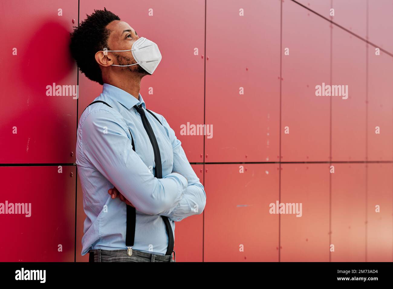 Businessman with a face mask looking up while leaning on a wall with his arms crossed. Stock Photo