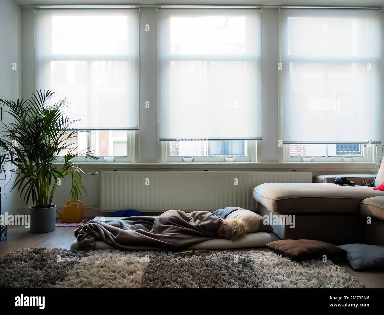 A teenage boy is sleeping on a matress in an improvised bed on the floor of a living room. Stock Photo