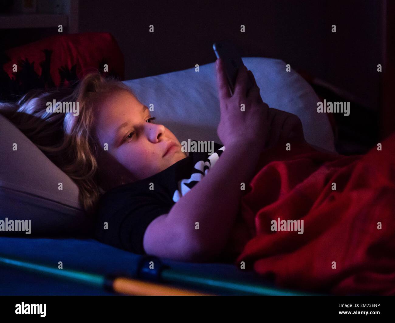 A teenage boy is lying in bed holding his smartphone before falling asleep. Smartphone display is illuminating boy's face. Caucasian ethnicity. Stock Photo