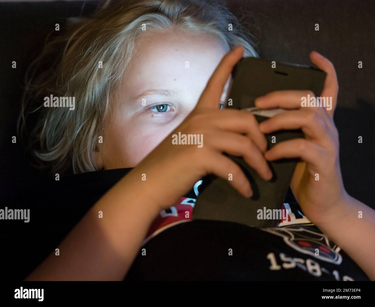 A teenage boy is lying in bed holding his smartphone before falling asleep. Smartphone display is illuminating boy's face. Caucasian ethnicity. Stock Photo