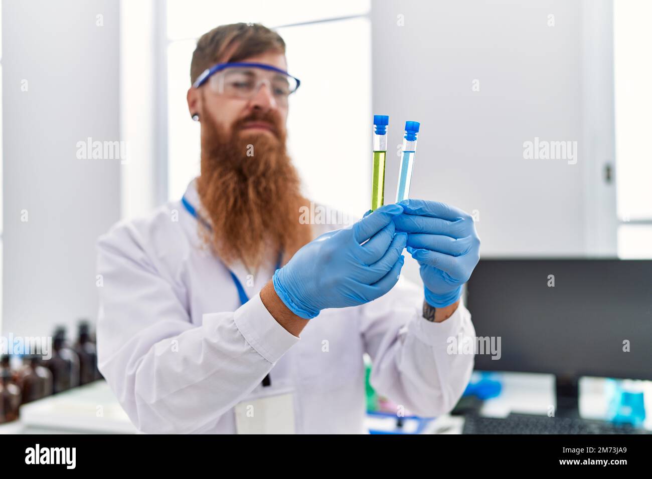 Young redhead man wearing scientist uniform holding test tubes at ...