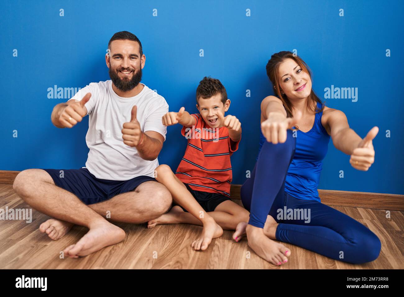 Family of three sitting on the floor at home approving doing positive gesture with hand, thumbs up smiling and happy for success. winner gesture. Stock Photo