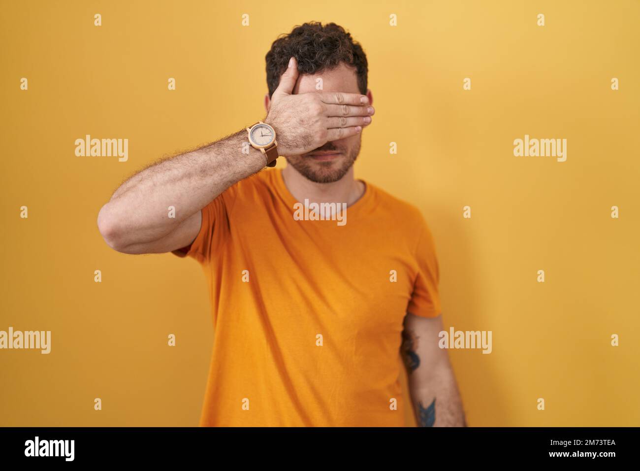 Young hispanic man standing over yellow background covering eyes with ...
