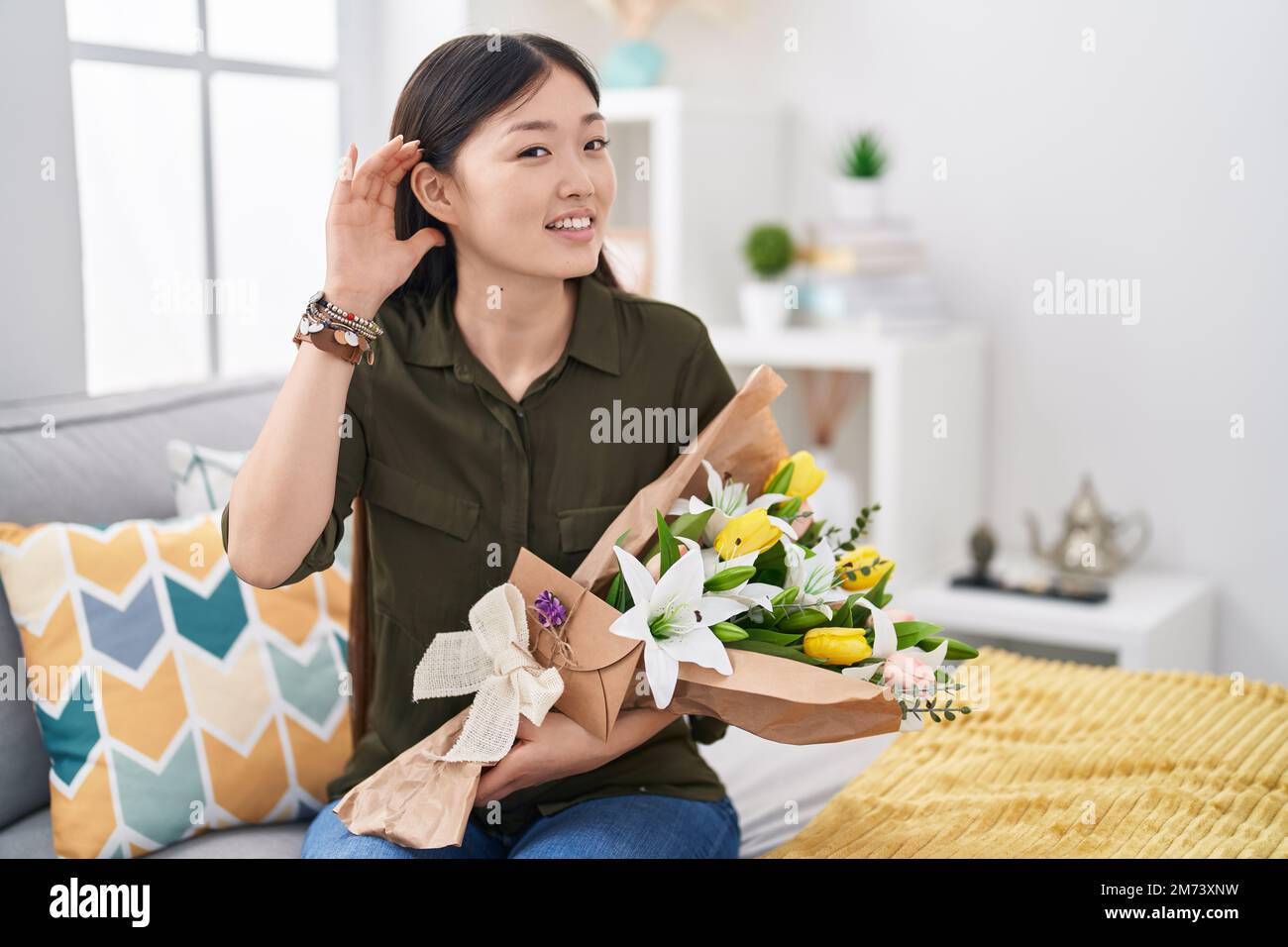 Chinese young woman holding bouquet of white flowers smiling with hand over ear listening an hearing to rumor or gossip. deafness concept. Stock Photo