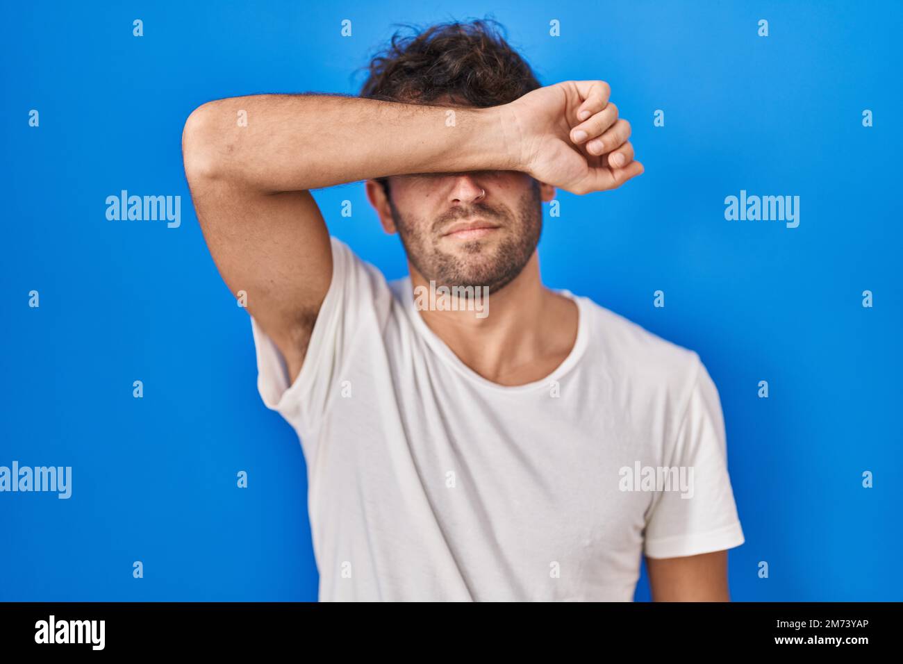 Hispanic young man standing over blue background covering eyes with arm ...