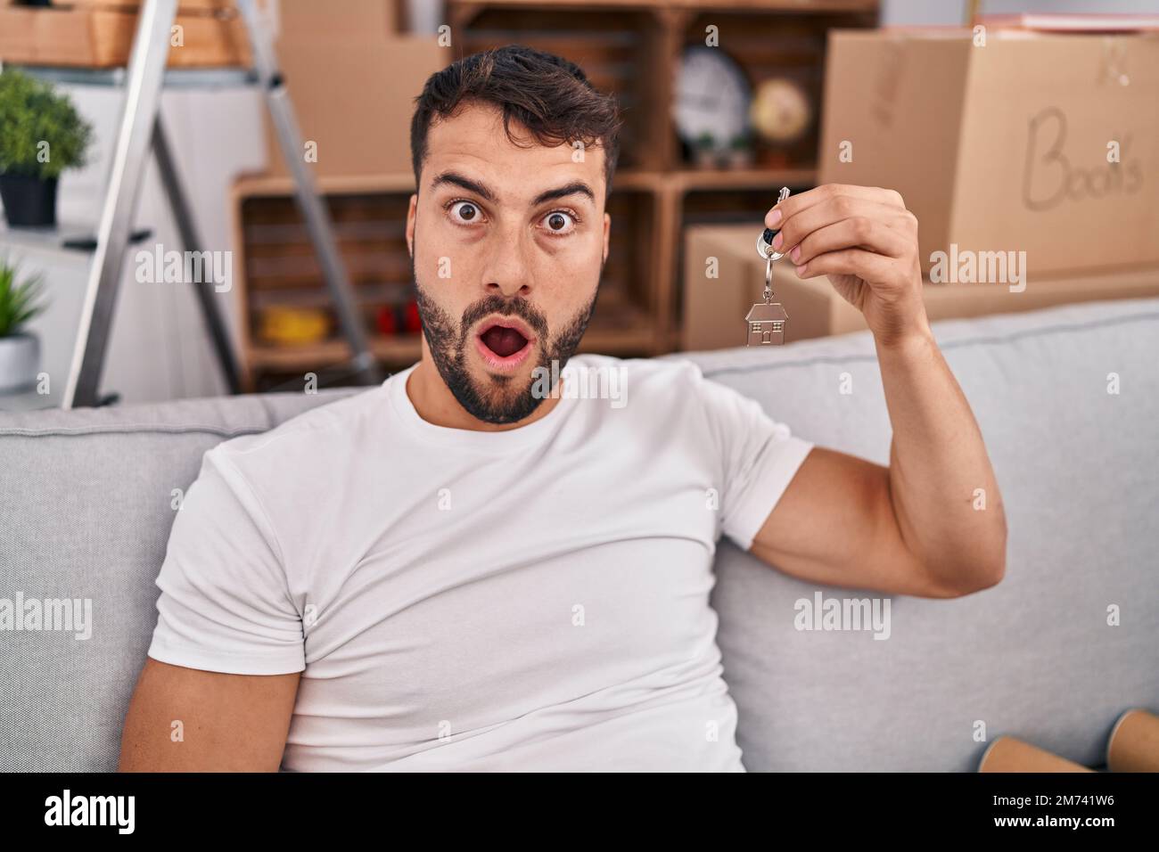 Handsome hispanic man holding keys of new home scared and amazed with open mouth for surprise