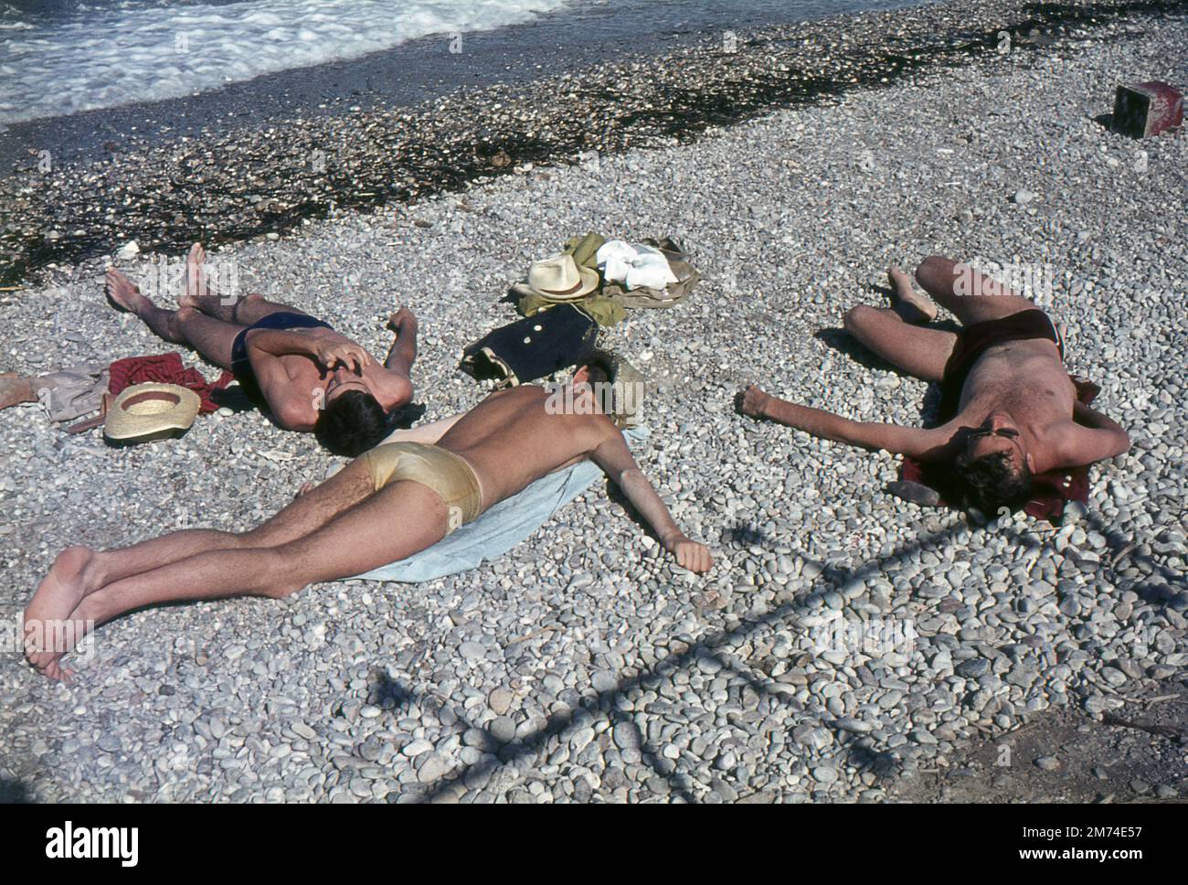 Xylokastro, Greece. August 1963. Three young men wearing swimming trunks are sunbathing on the shingle beach at the seaside town of Xylokastro in Corinthia in the Peloponnese, Greece. The beach showers are casting an interesting shadow on the pebbles. Stock Photo