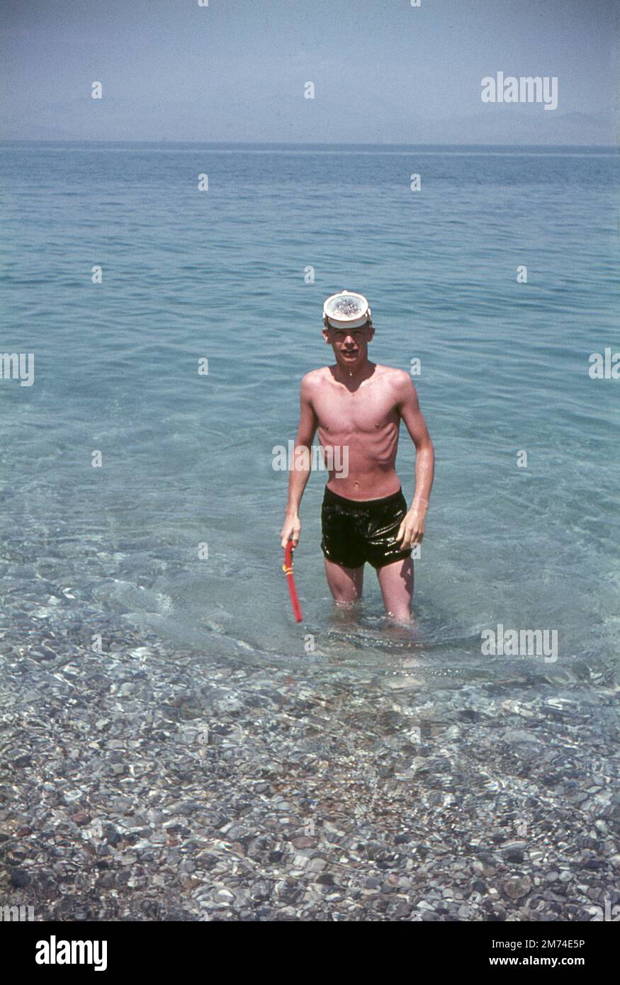 Xylokastro, Greece. August 1963. A young man returning to the beach after snorkelling in the Gulf of Corinth at the seaside town of Xylokastro in Corinthia in the Peloponnese, Greece. He has a scuba diving mask on his forehead and is holding a snorkel in his hand. The hills/mountains on the Greek mainland are visible on the horizon. Stock Photo