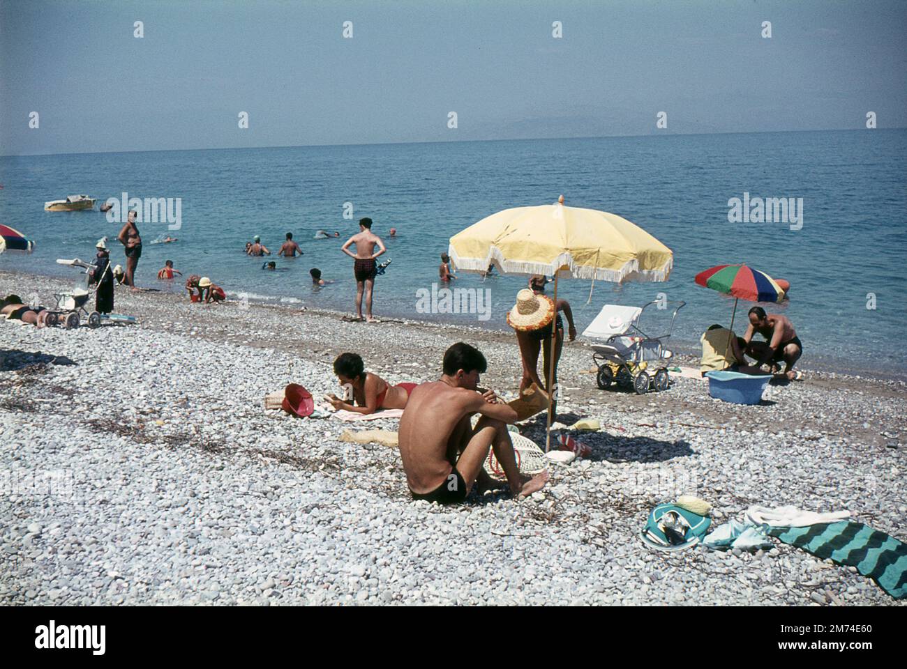 Xylokastro, Greece. August 1963. Tourists and local families relaxing on the beach at the seaside town of Xylokastro in Corinthia in the Peloponnese, Greece. The visitors are sunbathing and sheltering under parasols, while others are swimming in the Gulf of Corinth. The hills/mountains on the Greek mainland are visible on the horizon. Stock Photo