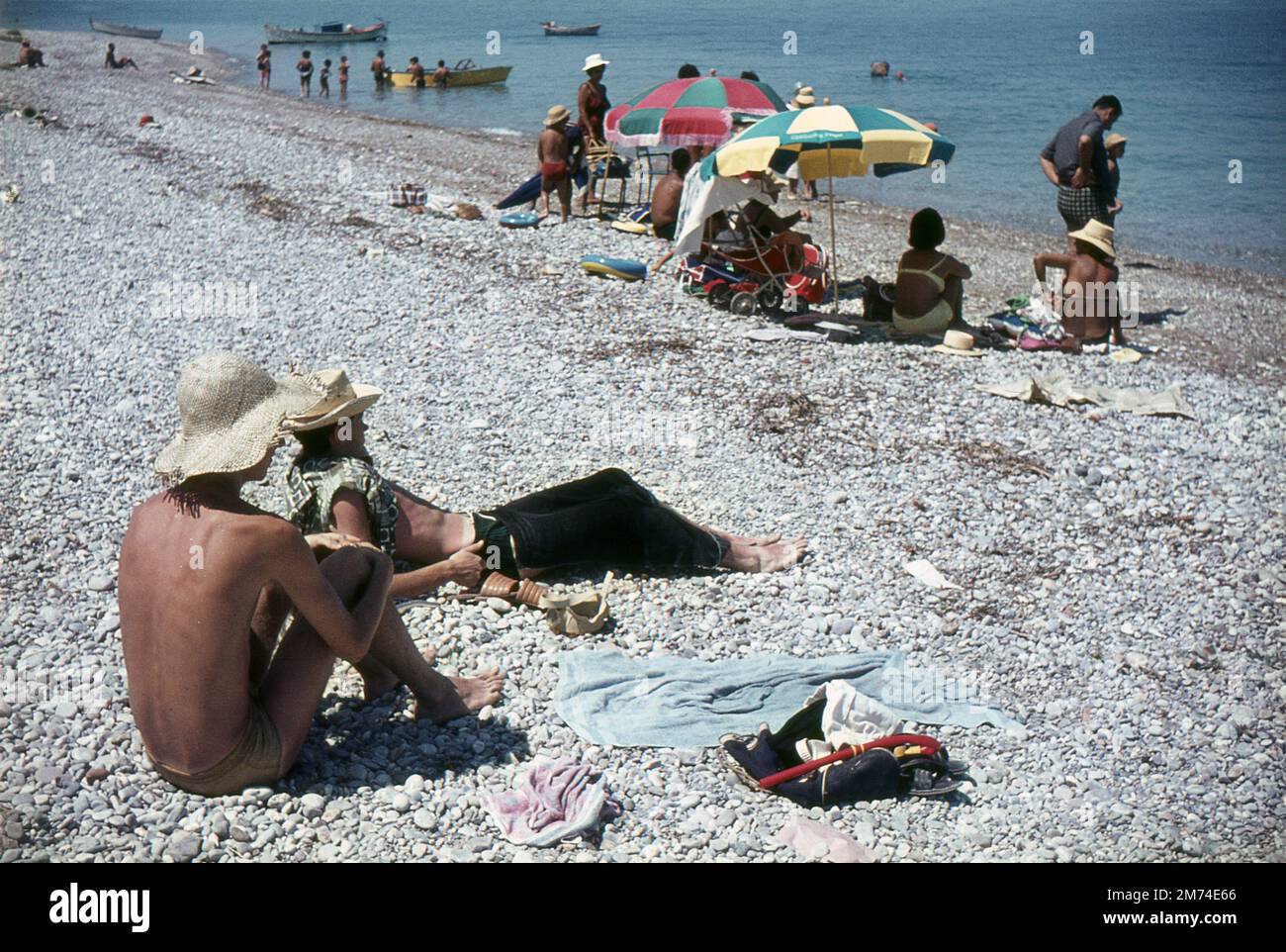 Xylokastro, Greece. August 1963. Two young men, other tourists and local families are relaxing on the beach at the seaside town of Xylokastro in Corinthia in the Peloponnese, Greece. The visitors are sunbathing, some sheltering under parasols, whilst others are swimming in the Gulf of Corinth. Various boats are moored just off the beach. Stock Photo